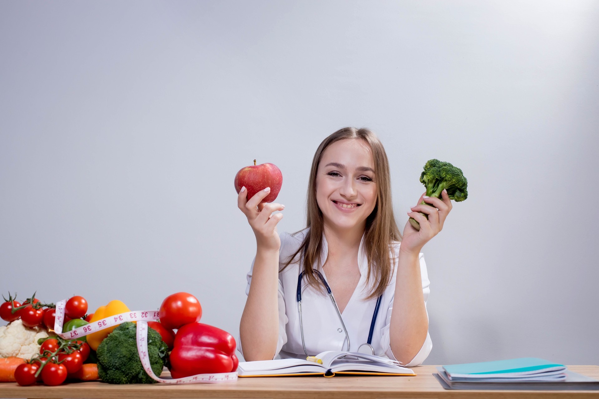 Positive young female nutritionist sitting at the table and holding broccoli and an apple in her hands. Concept of healthy eating